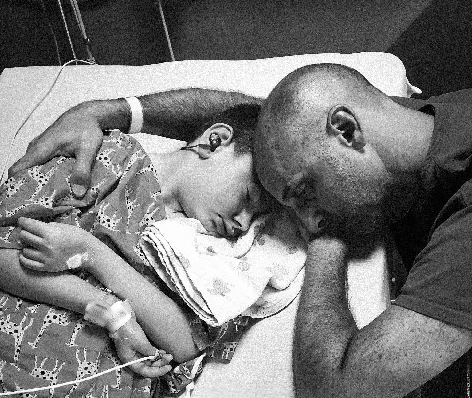 A sleeping disabled boy with hospital tags, embraced by his caregiving father